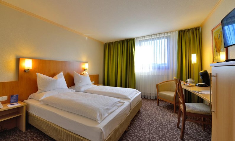 Hotelzimmer mit Teppichboden, Doppelbett, großem Fenster, Stuhl, Tisch, Sessel, gemütlich beleuchtet, beige und grüne Vorhänge